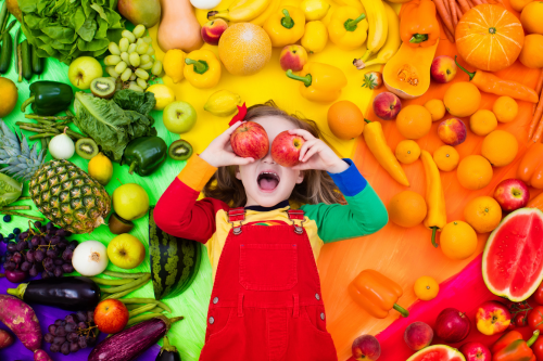 Niña sonriendo y sosteniendo manzanas sobre sus ojos acostada sobre una variedad de frutas y verduras coloridas y saludables