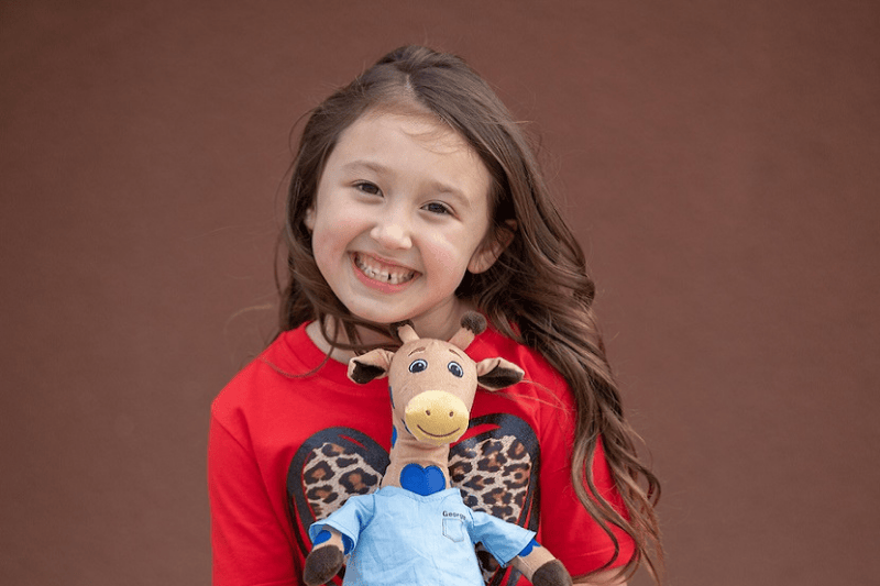 Una niña sonríe mientras sostiene un juguete de peluche de George la jirafa
