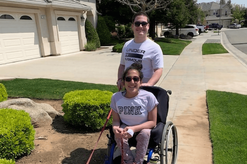 A teenage boy and a teenage girl in a wheelchair take a walk in their neighborhood