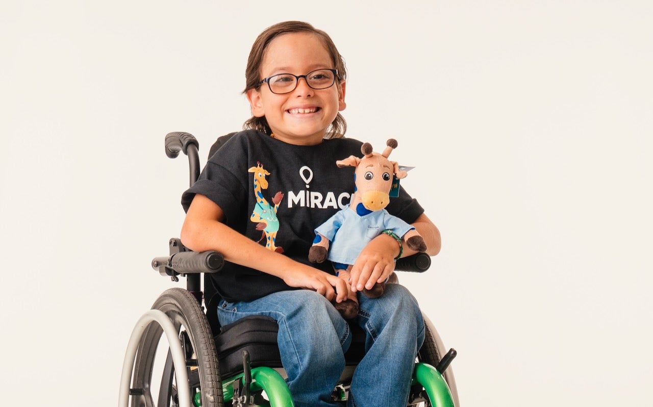a little boy in a wheelchair smiles while holding a George the Giraffe stuffed animal