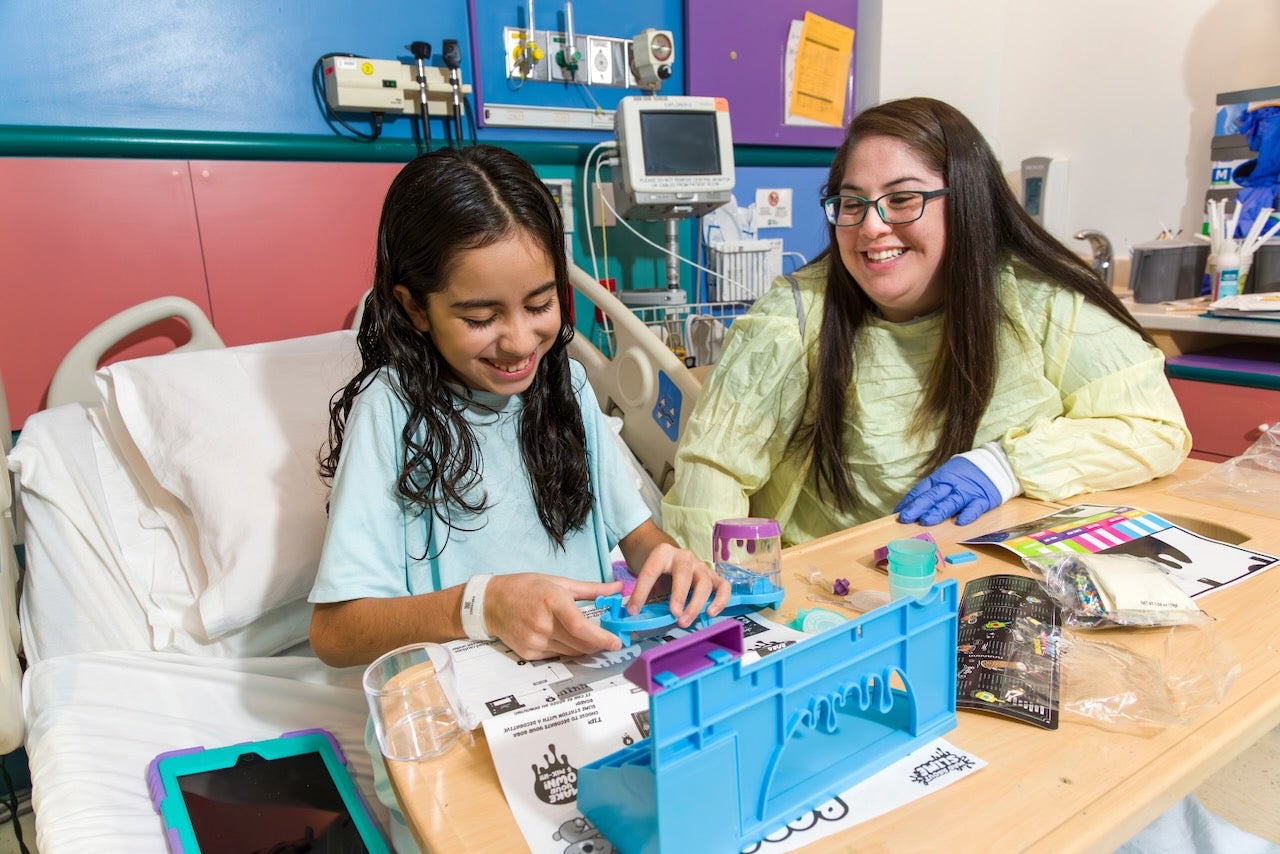Una niña juega con juguetes en la mesita de su cama de hospital junto con un especialista en vida hospitalaria infantil