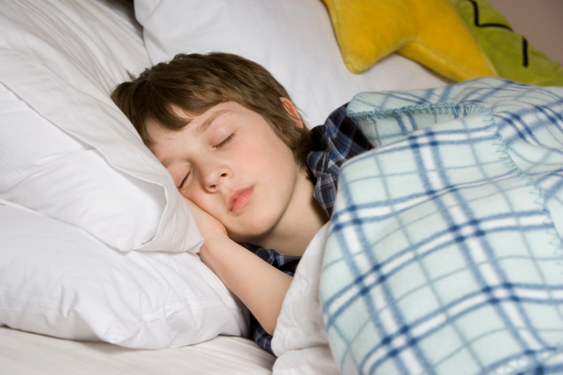 A young boy sleeps in bed under a blanket.
