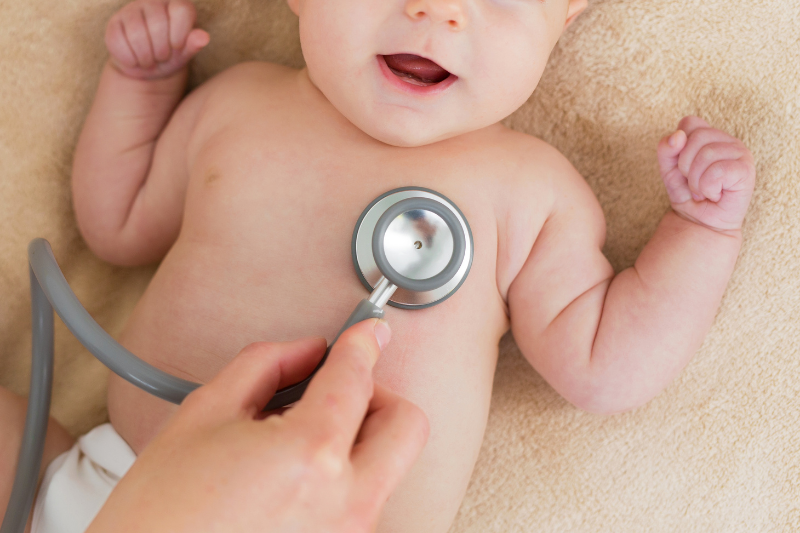 A baby lies on a blanket checked by a stethoscope.