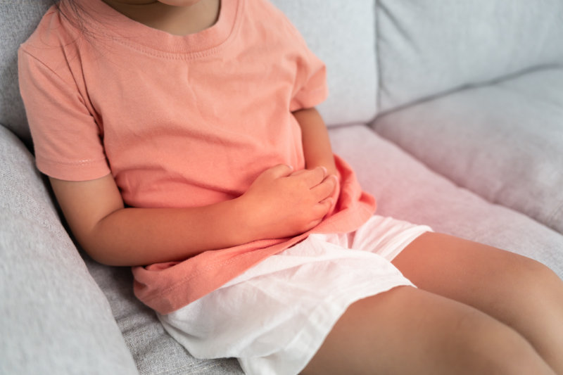 A young girl sits on a couch with stomach pain.